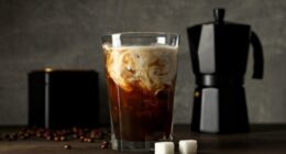 Glass with iced coffee drink, coffee maker, coffee bean and sugar on gray background